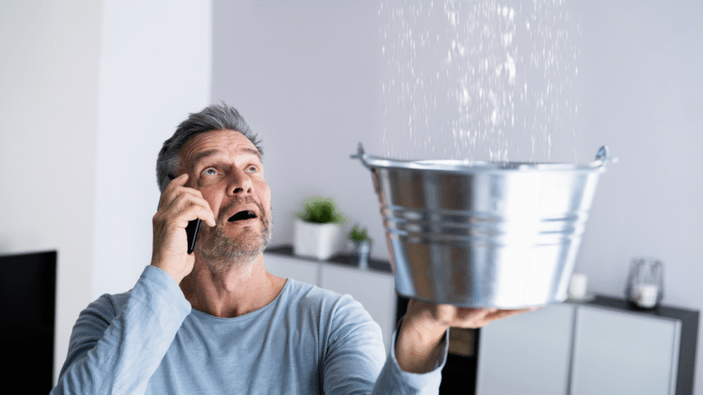 A man holding a bucket to catch water from a roof leak while talking on the phone to schedule an emergency roofing service.