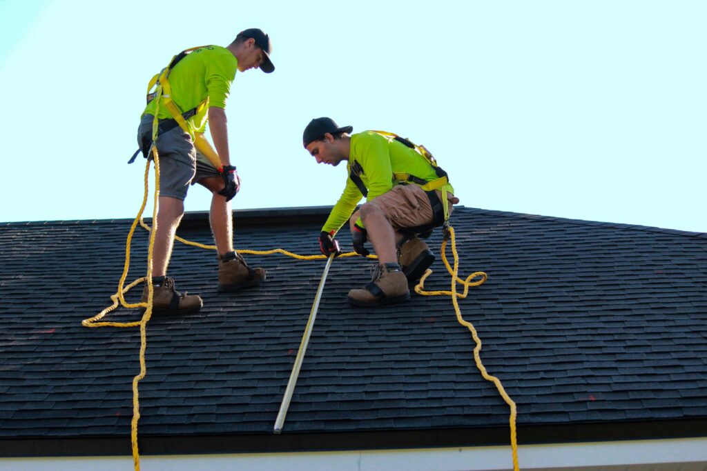 Two professional roofers inspecting a black shingled roof with a measuring tool.