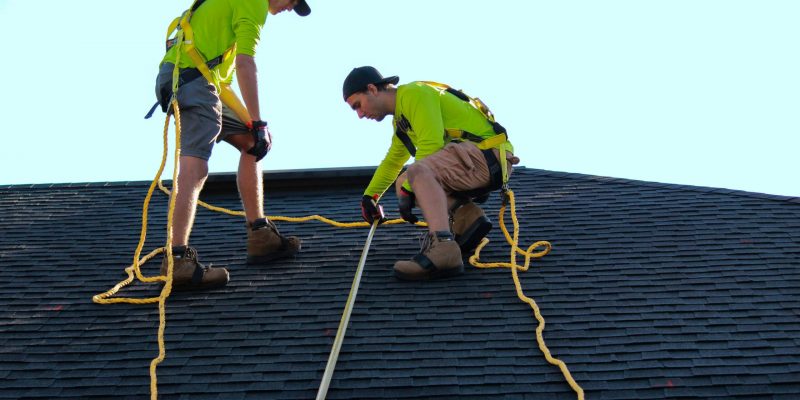 Two professional roofers inspecting a black shingled roof with a measuring tool.